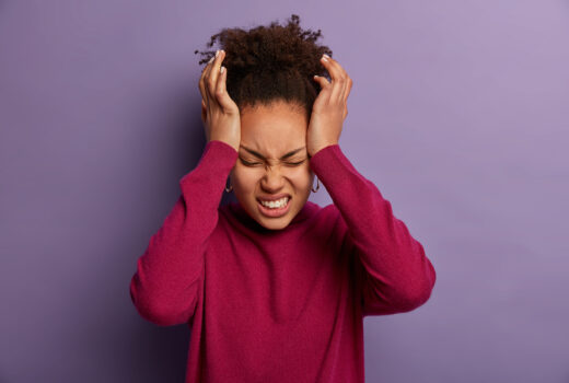 Photo of dissatisfied woman keeps hands on temple, suffers from unbearable headache, clenches teeth with pain, being exhausted after work, wears burgundy turtleneck, isolated on purple wall. Photo of dissatisfied woman keeps hands on temple, suffers from unbearable headache, clenches teeth with pain, being exhausted after work, wears burgundy turtleneck, isolated on purple wall.